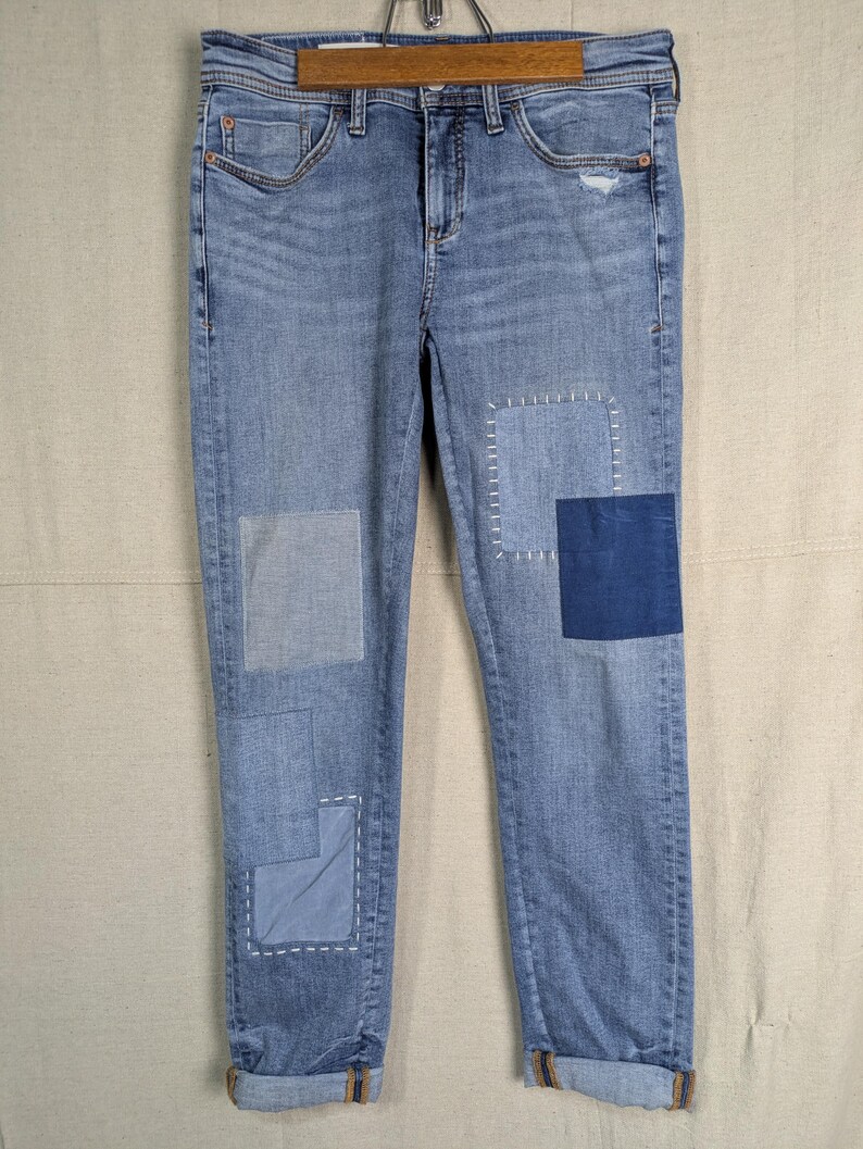 May include: Light blue denim jeans with patches of varying shades of blue. The jeans have a distressed look with a small rip on the right knee. The cuffs are rolled up, revealing a contrasting fabric.