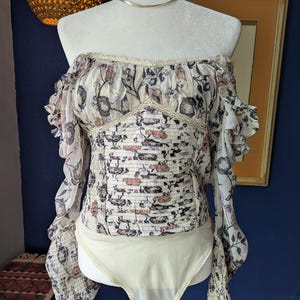 May include: Off-the-shoulder bodysuit with a floral print in cream, pink, and blue. The top has long, ruffled sleeves and a fitted bodice. The bottom is a solid cream color.