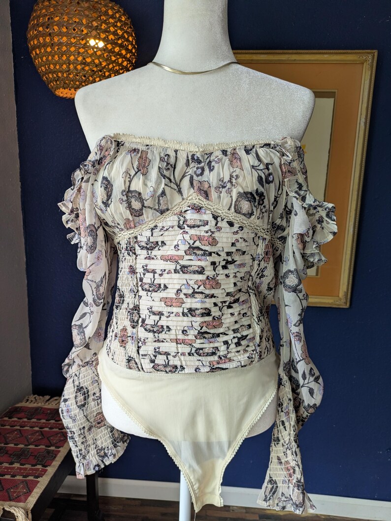 May include: Off-the-shoulder bodysuit with a floral print in cream, pink, and blue. The top has long, ruffled sleeves and a fitted bodice. The bottom is a solid cream color.