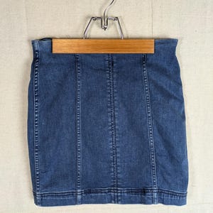 May include: A dark wash denim skirt hangs on a wooden hanger. The skirt has vertical seams and a classic, fitted design. The skirt is a mini-length and appears to be made of a sturdy denim material.