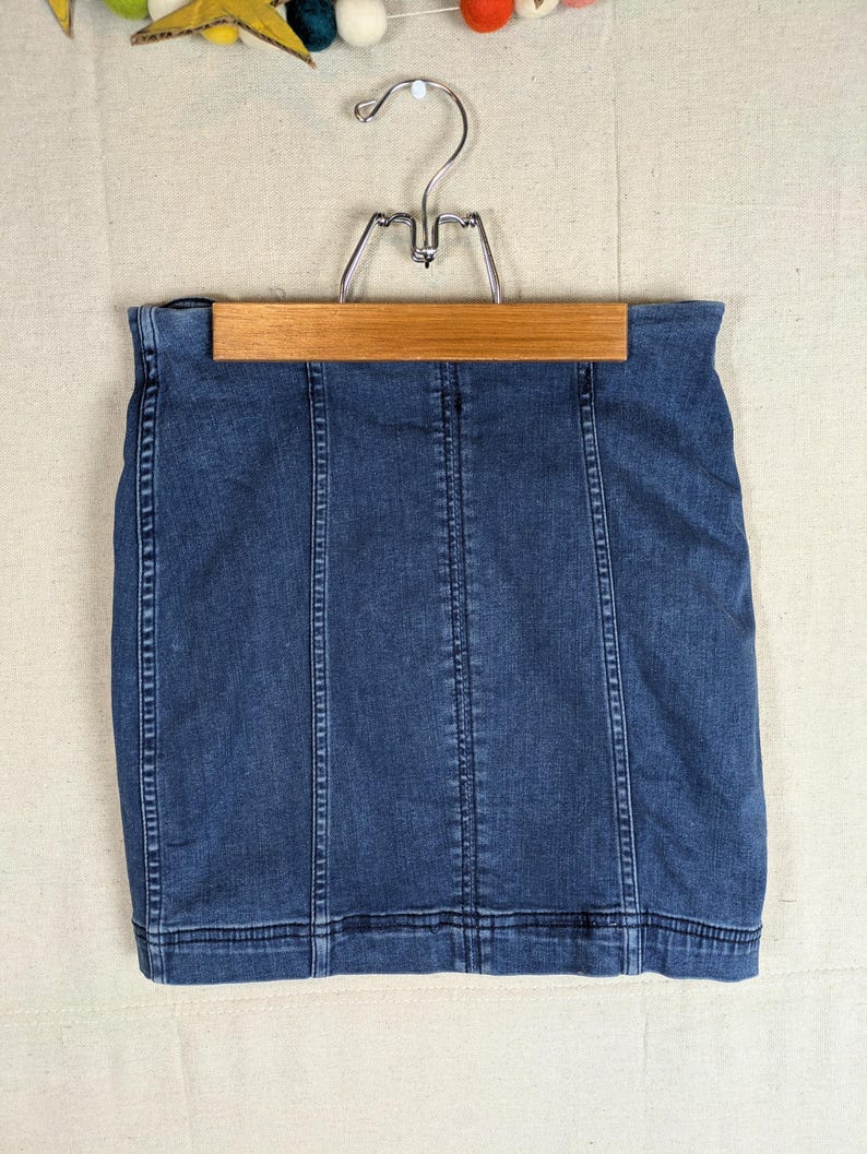 May include: A dark wash denim skirt hangs on a wooden hanger. The skirt has vertical seams and a classic, fitted design. The skirt is a mini-length and appears to be made of a sturdy denim material.