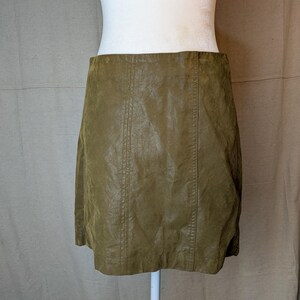 May include: Olive green faux leather skirt. The skirt has a slight A-line shape and features vertical seams down the front and back. The skirt is displayed on a white mannequin.