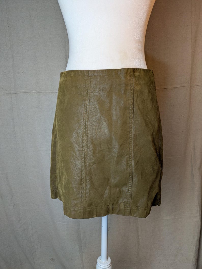 May include: Olive green faux leather skirt. The skirt has a slight A-line shape and features vertical seams down the front and back. The skirt is displayed on a white mannequin.