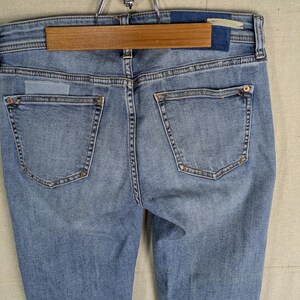 May include: Light blue denim jeans with a classic five-pocket design. The jeans are hanging on a wooden hanger. The back pockets have decorative stitching and rivets. The denim has a faded wash.