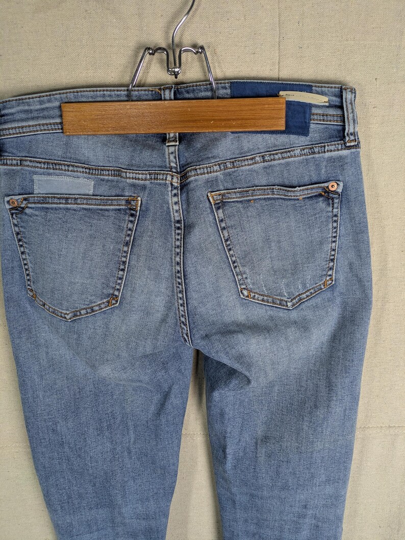 May include: Light blue denim jeans with a classic five-pocket design. The jeans are hanging on a wooden hanger. The back pockets have decorative stitching and rivets. The denim has a faded wash.