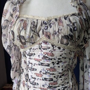 May include: A cream-colored top with a floral pattern in shades of pink, purple, and black. The top features long, sheer sleeves and a square neckline with delicate lace trim. The bodice is ruched, creating a fitted silhouette.