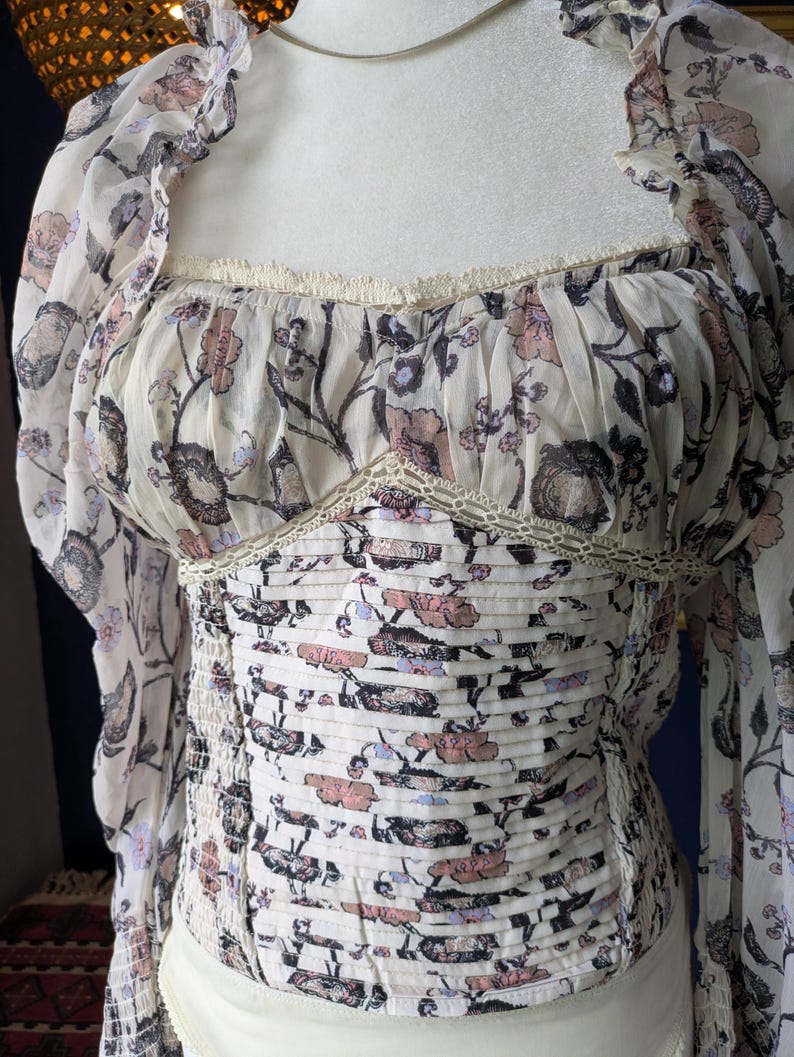 May include: A cream-colored top with a floral pattern in shades of pink, purple, and black. The top features long, sheer sleeves and a square neckline with delicate lace trim. The bodice is ruched, creating a fitted silhouette.