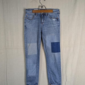 May include: Light blue denim jeans with patches of varying shades of blue. The jeans are cuffed at the ankles and hang from a wooden hanger. The patches are square and rectangular, with visible stitching.