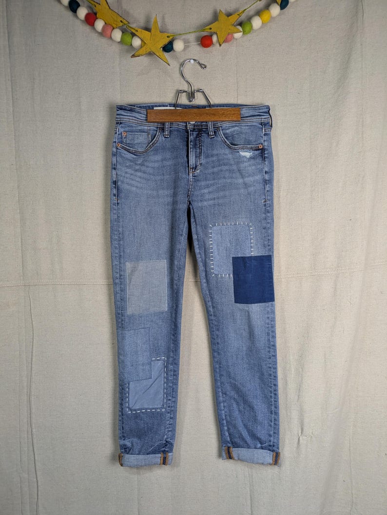 May include: Light blue denim jeans with patches of varying shades of blue. The jeans are cuffed at the ankles and hang from a wooden hanger. The patches are square and rectangular, with visible stitching.