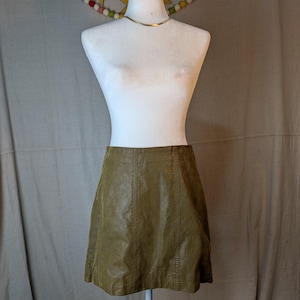 May include: A-line mini skirt in a muted olive green hue. The skirt is made of a faux leather material and features vertical seam detailing. The skirt is displayed on a white mannequin with a gold-colored necklace.
