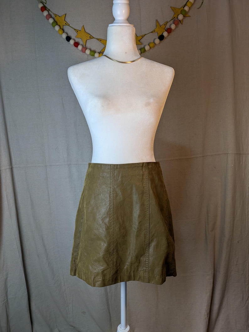 May include: A-line mini skirt in a muted olive green hue. The skirt is made of a faux leather material and features vertical seam detailing. The skirt is displayed on a white mannequin with a gold-colored necklace.