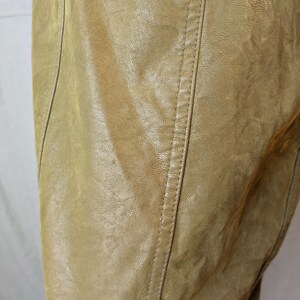 May include: Close-up of a tan leather garment, possibly a jacket or coat. The leather has a textured appearance with visible grain and subtle color variations. A vertical seam with detailed stitching runs down the center, adding a tailored element to the design.