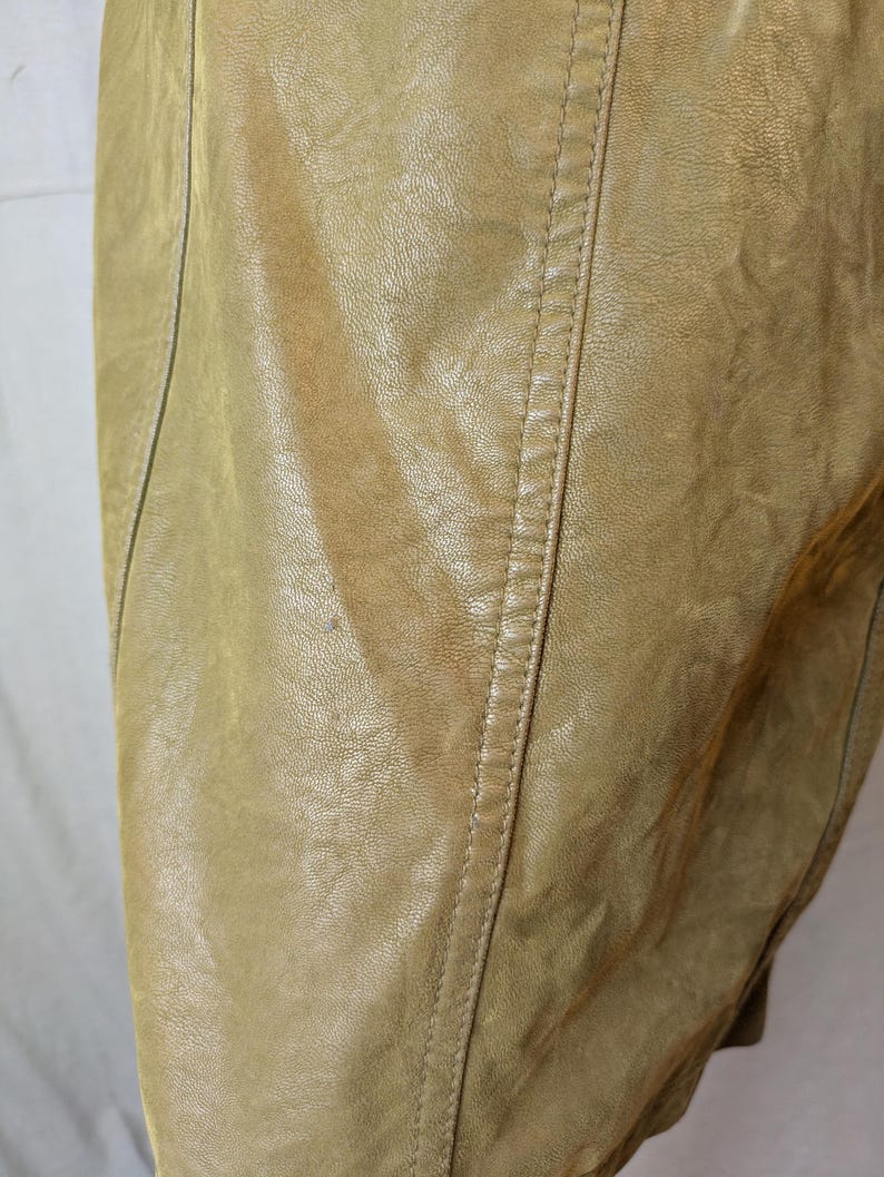 May include: Close-up of a tan leather garment, possibly a jacket or coat. The leather has a textured appearance with visible grain and subtle color variations. A vertical seam with detailed stitching runs down the center, adding a tailored element to the design.