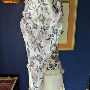 May include: A cream-colored top with a floral pattern in shades of pink, gray, and black. The top has a sheer, flowing overlay and a fitted bodice. The design includes a ruffled neckline and sleeves, creating a bohemian aesthetic.