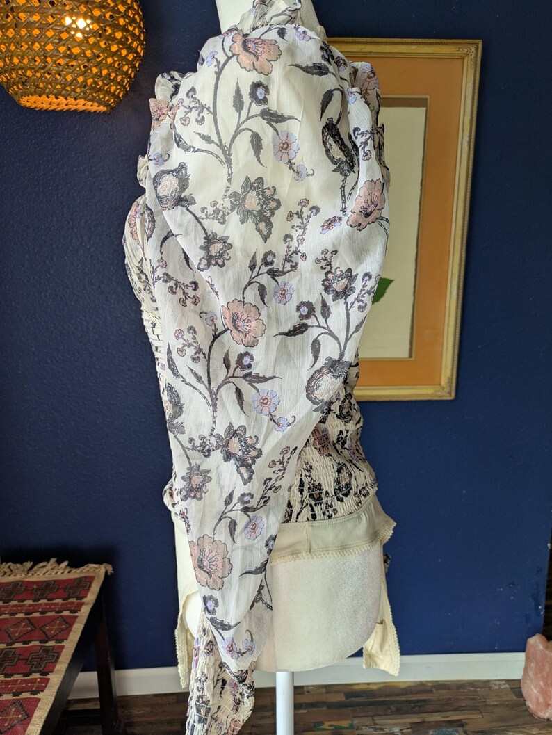 May include: A cream-colored top with a floral pattern in shades of pink, gray, and black. The top has a sheer, flowing overlay and a fitted bodice. The design includes a ruffled neckline and sleeves, creating a bohemian aesthetic.