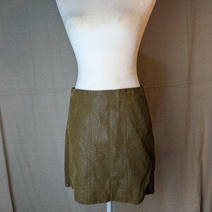 May include: Olive green faux leather mini skirt. The skirt is displayed on a white mannequin. The skirt has vertical seams down the front and back. The skirt is a classic A-line shape.