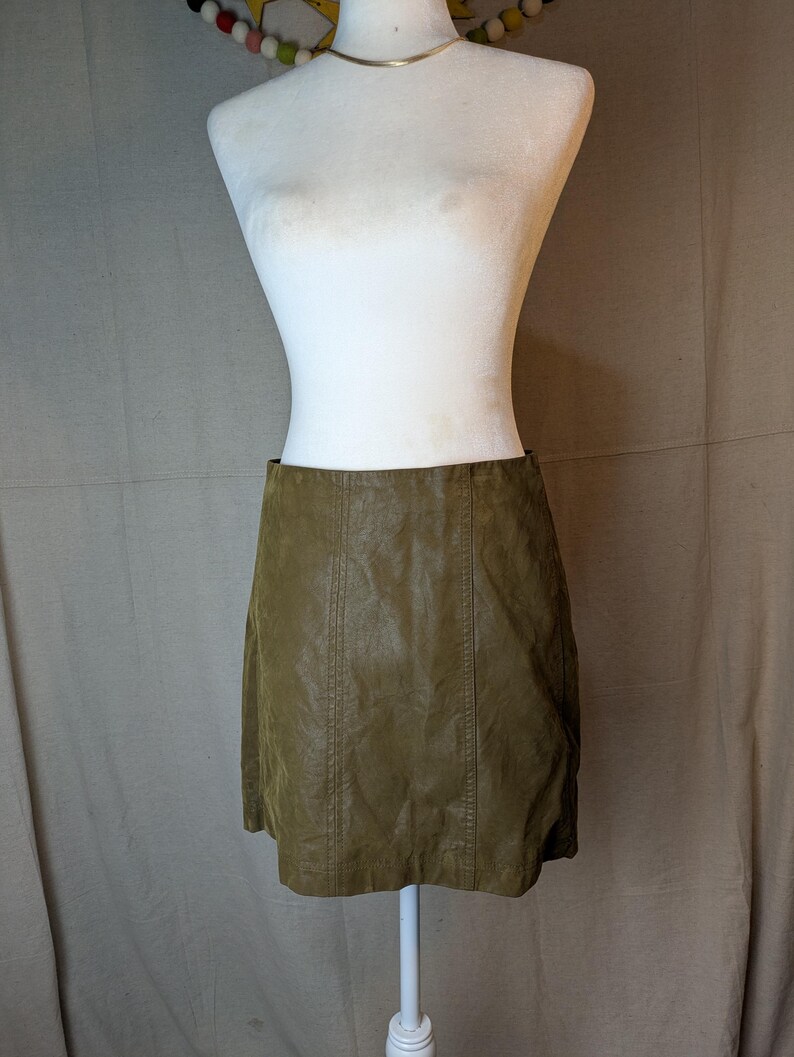 May include: Olive green faux leather mini skirt. The skirt is displayed on a white mannequin. The skirt has vertical seams down the front and back. The skirt is a classic A-line shape.