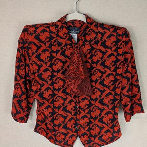 May include: A black blouse with a vibrant red floral pattern, featuring three-quarter length sleeves, a pointed collar, and a front button closure. The garment includes a decorative neck tie with a striped accent and a Carole Little brand label.