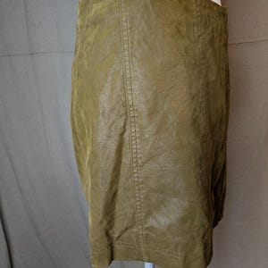 May include: Olive green faux leather skirt. The skirt has a straight silhouette and a high waist. Vertical seams run down the front and back. The skirt is displayed on a white mannequin.