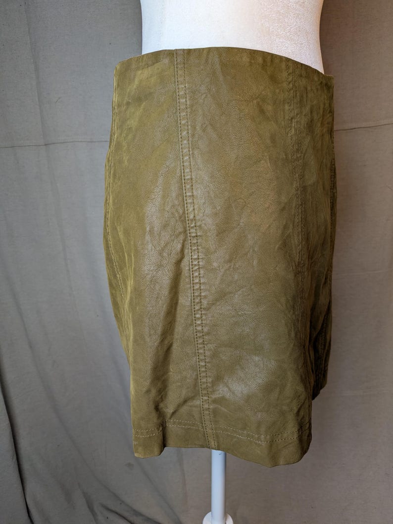 May include: Olive green faux leather skirt. The skirt has a straight silhouette and a high waist. Vertical seams run down the front and back. The skirt is displayed on a white mannequin.