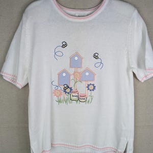 May include: A white short-sleeved knit sweater featuring a pink-trimmed crew neckline and hem. The front displays an embroidered design of three birdhouses, two bees, flowers, and two jars labeled honey, with small white buttons on the birdhouses.