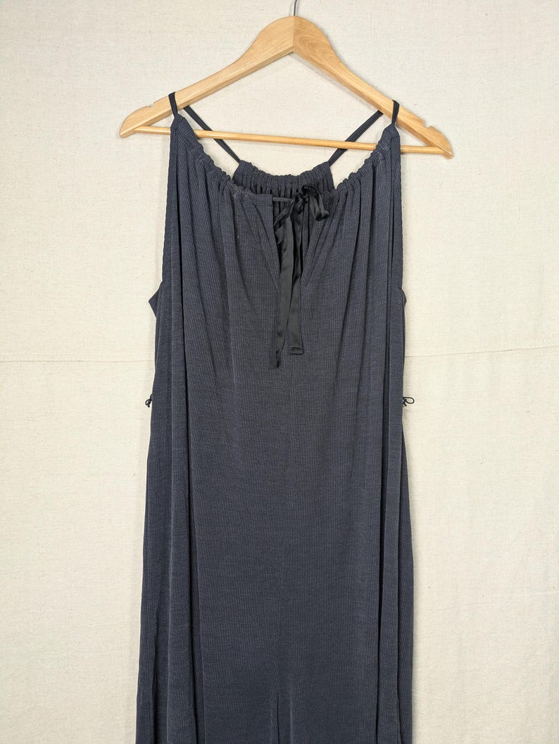 May include: A dark gray, sleeveless dress with a halter neckline and a keyhole detail. The dress has a flowing, ribbed texture and is hanging on a wooden hanger. A black ribbon is tied at the neckline.