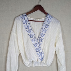 May include: White long-sleeved blouse with a V-neckline and blue floral embroidery along the edges. The blouse has a wrap-style design and is hanging on a wooden hanger. The fabric has a subtle striped texture.