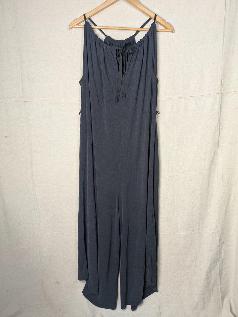 May include: A dark gray jumpsuit with a halter neckline and a tie detail at the chest. The jumpsuit has wide legs and is hanging on a wooden hanger.