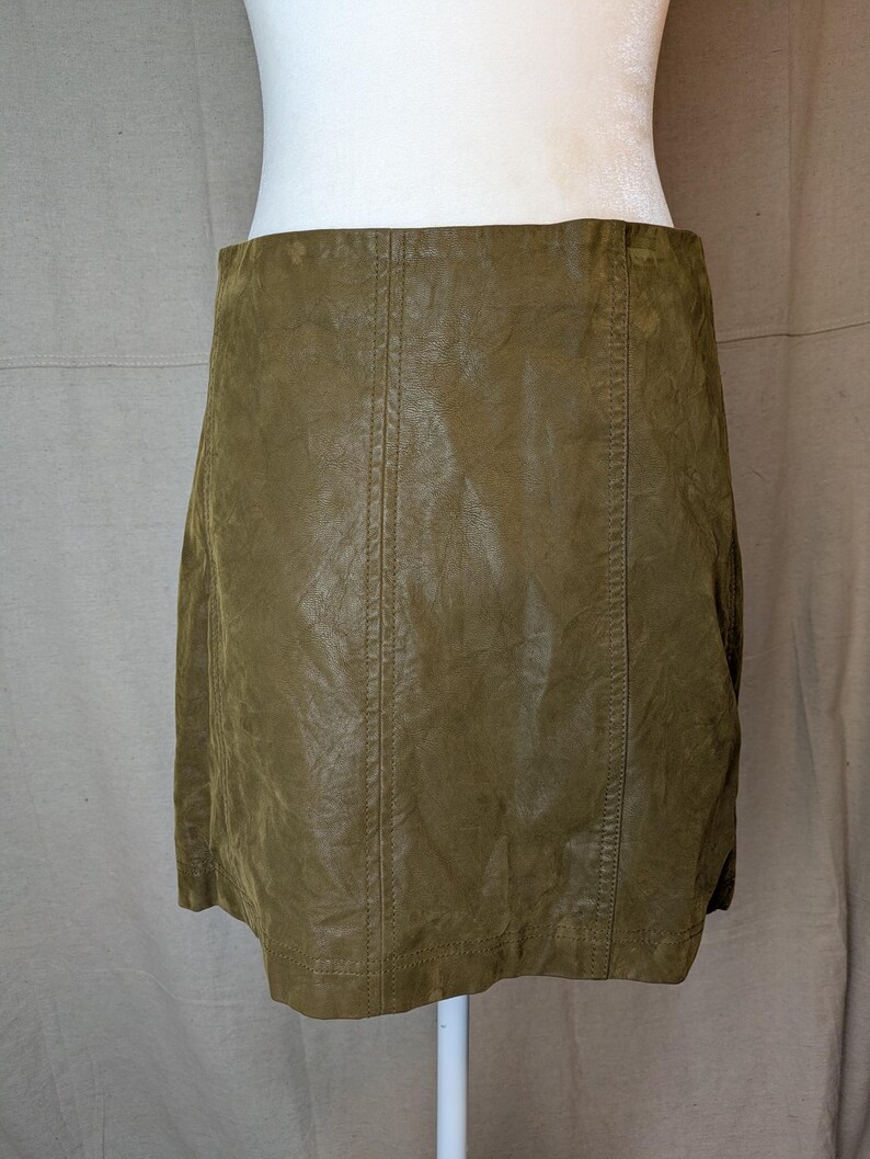 May include: Olive green mini skirt made of a textured material. The skirt has vertical seams down the front and back. The skirt is displayed on a white mannequin.