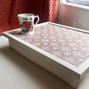 May include: A white tray with a red, yellow and grey patterned design. The tray has a white frame and a soft, padded base. A white mug with a red apple design is sitting on the tray.