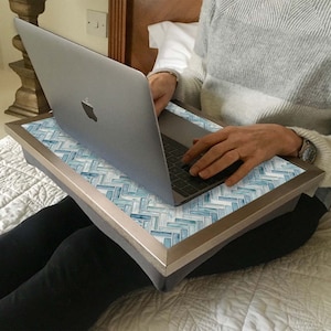May include: A blue and white tiled laptop tray with a silver frame. The tray is sitting on a bed and a person is using a laptop on the tray.