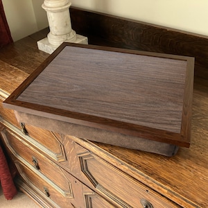 May include: A brown wooden tray with a dark brown woodgrain finish and a brown suede cushion. The tray is sitting on a wooden dresser.