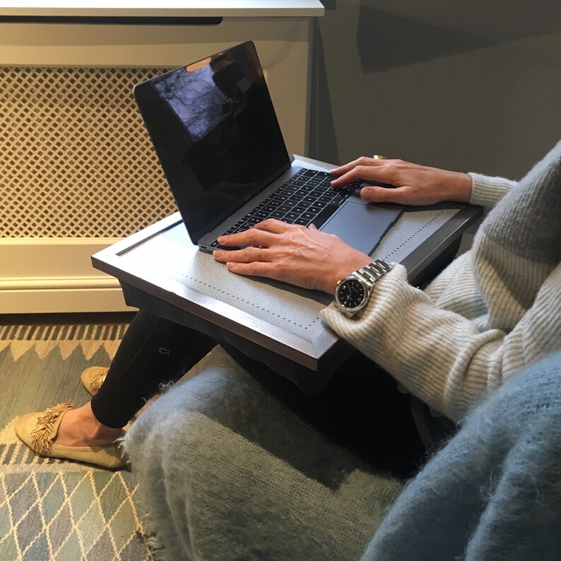 May include: A person using a laptop computer on a gray wooden lap desk. The desk has a textured surface and a raised edge. The person is wearing a white sweater and a watch.