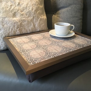May include: A brown tray with a padded base and a decorative tile design. The tray is sitting on a grey sofa with a white faux fur cushion and a grey cushion. A white coffee cup with a brown handle is sitting on a white saucer on the tray.