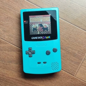 Könnte beinhalten: Eine türkisfarbene Game Boy Color Handheld-Spielkonsole mit einer Donkey Kong Country Spielkassette. Der Bildschirm zeigt den Spieltitel und einen Charakter aus dem Spiel.