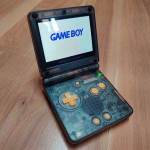 May include: A translucent black Game Boy Advance SP with a clear green power light. The screen displays the text "GAME BOY" in blue. The buttons are yellow and the device is on a wooden surface.