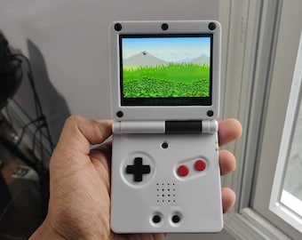 Puede incluir: Una consola de juegos portátil blanca con una pantalla negra que muestra un paisaje verde. La consola tiene un pad direccional negro, dos botones rojos y el texto "GAME BOY ADVANCE SP" en la pantalla.