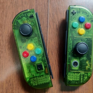 Puede incluir: Dos mandos de juego verdes transparentes con joysticks negros y botones de colores. Los mandos tienen un diseño transparente, que revela los componentes internos. Los botones son rojos, amarillos, azules y verdes. Diseñado para juegos.