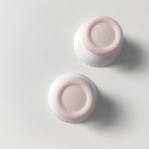 May include: Two white, round cosmetic containers with a light pink, textured surface. The containers are set against a white background, creating a clean and minimalist aesthetic. The design suggests a focus on skincare or beauty products.