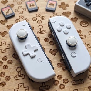 May include: Two white and gray game controllers with a D-pad and buttons, along with several game cartridges. The controllers are on a wooden surface decorated with video game-themed icons, including hearts, directional pads, and controller shapes.