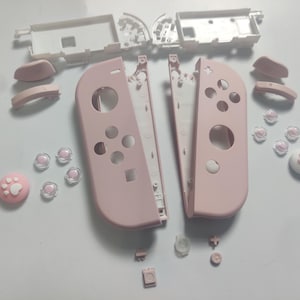 Op de afbeelding: Onderdelen van een roze gamecontroller, gedemonteerd, waaronder Joy-Con-schelpen, knoppen en een joystick in de vorm van een poot. De onderdelen zijn gerangschikt op een wit oppervlak, wat de interne structuur en aanpassingsmogelijkheden benadrukt.