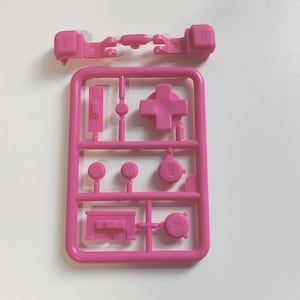 May include: Pink plastic mold with various shapes for video game controller buttons. The mold includes a plus sign, a circle, a square, and a triangle.