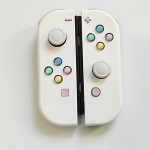 以下が含まれることがあります： ピンクのアクセントとカラフルなボタンが付いた白いゲームコントローラーのペア。各コントローラーには、グレーのジョイスティックと、青、緑、黄色、ピンクなどのマルチカラーボタンのセットがあります。コントローラーはゲーム用に設計されています。