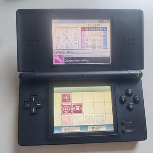 May include: A matte black handheld gaming console with dual screens. The top screen displays a clock and calendar, while the bottom screen shows a grid menu with icons for settings and system options. The device features a directional pad and buttons.