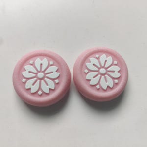 May include: Two pink, circular decorative items with a white floral design in the center. The floral design is a stylized flower with multiple petals. The items are set against a white background.
