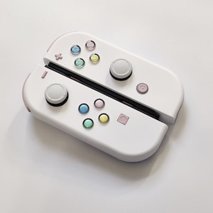 Puede incluir: Un mando Nintendo Switch Joy-Con blanco con detalles en rosa y botones de colores. El mando est&aacute; en posici&oacute;n cerrada, con las mitades izquierda y derecha del mando visibles.