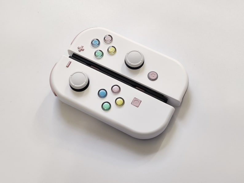 Puede incluir: Un mando Nintendo Switch Joy-Con blanco con detalles en rosa y botones de colores. El mando est&aacute; en posici&oacute;n cerrada, con las mitades izquierda y derecha del mando visibles.