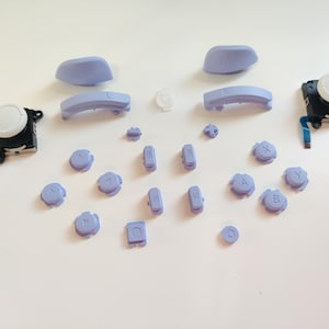May include: A set of replacement buttons for a video game controller. The buttons are purple and include the letters A, B, X, Y, and directional arrows. There are also two white analogue stick caps.