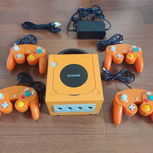 May include: An orange Nintendo GameCube console with four orange controllers. The console has a black screen with the text "Nintendo GameCube" printed on it.