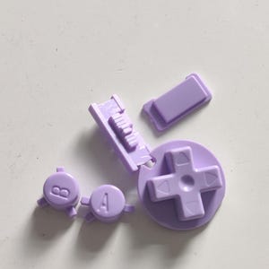 Peut inclure: Un ensemble de boutons de remplacement violet clair pour une manette de jeu, comprenant une croix directionnelle circulaire, deux boutons ronds marqués des lettres A et B, et un bouton rectangulaire, tous avec une finition en plastique mat.